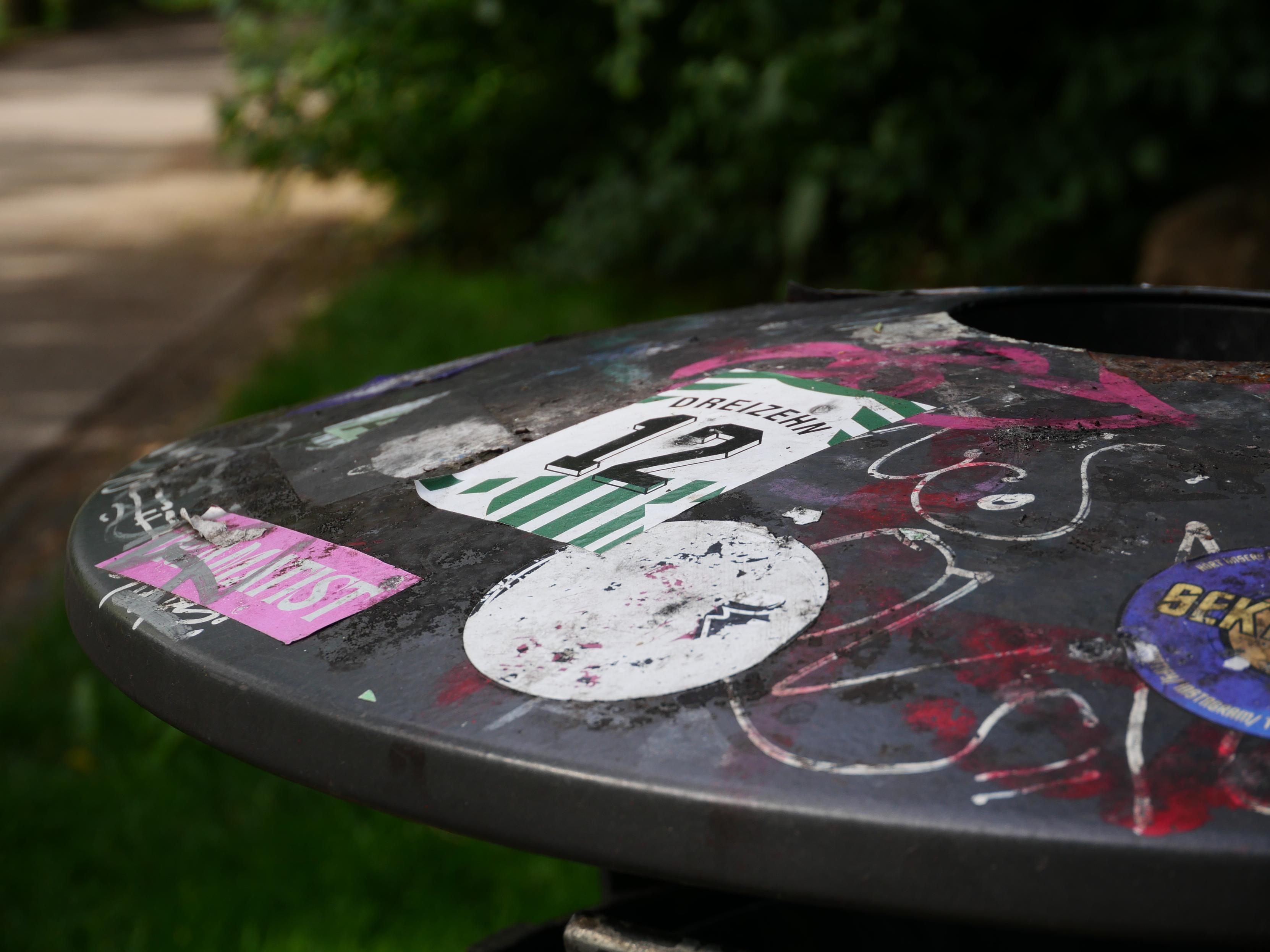 Nahaufnahme eines schwarzen Mülleimerdeckels mit zahlreichen Graffiti-Kritzeleien und Aufklebern, darunter ein grün-weißer Sticker mit der Nummer 12 und der Beschriftung 'Werder', ein pinker Sticker mit dem Wort 'Feminist', sowie ein blauer runder Aufkleber. Im Hintergrund unscharfe grüne Vegetation und ein Weg.